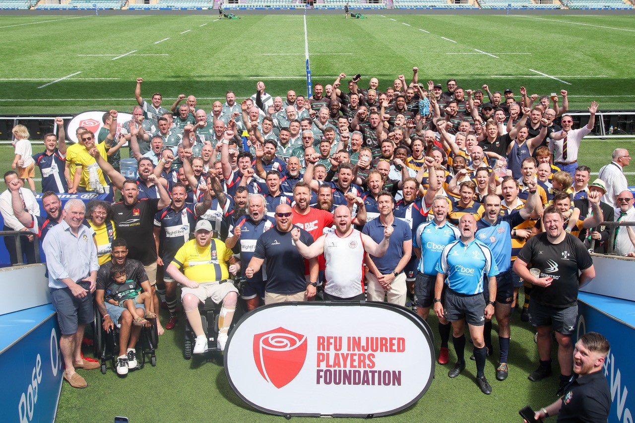 Image shows 100 male rugby players gathered together smiling with their hands up, whilst stood in the tunnel entrance to the Allianz Stadium, Twickenham pitch.
