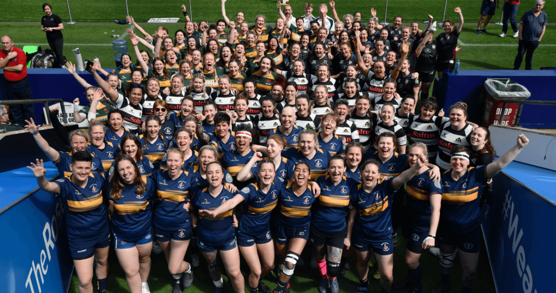 Image shows 100 female rugby players gathered together smiling with their hands up, whilst stood in the tunnel entrance to the Allianz Stadium, Twickenham pitch.