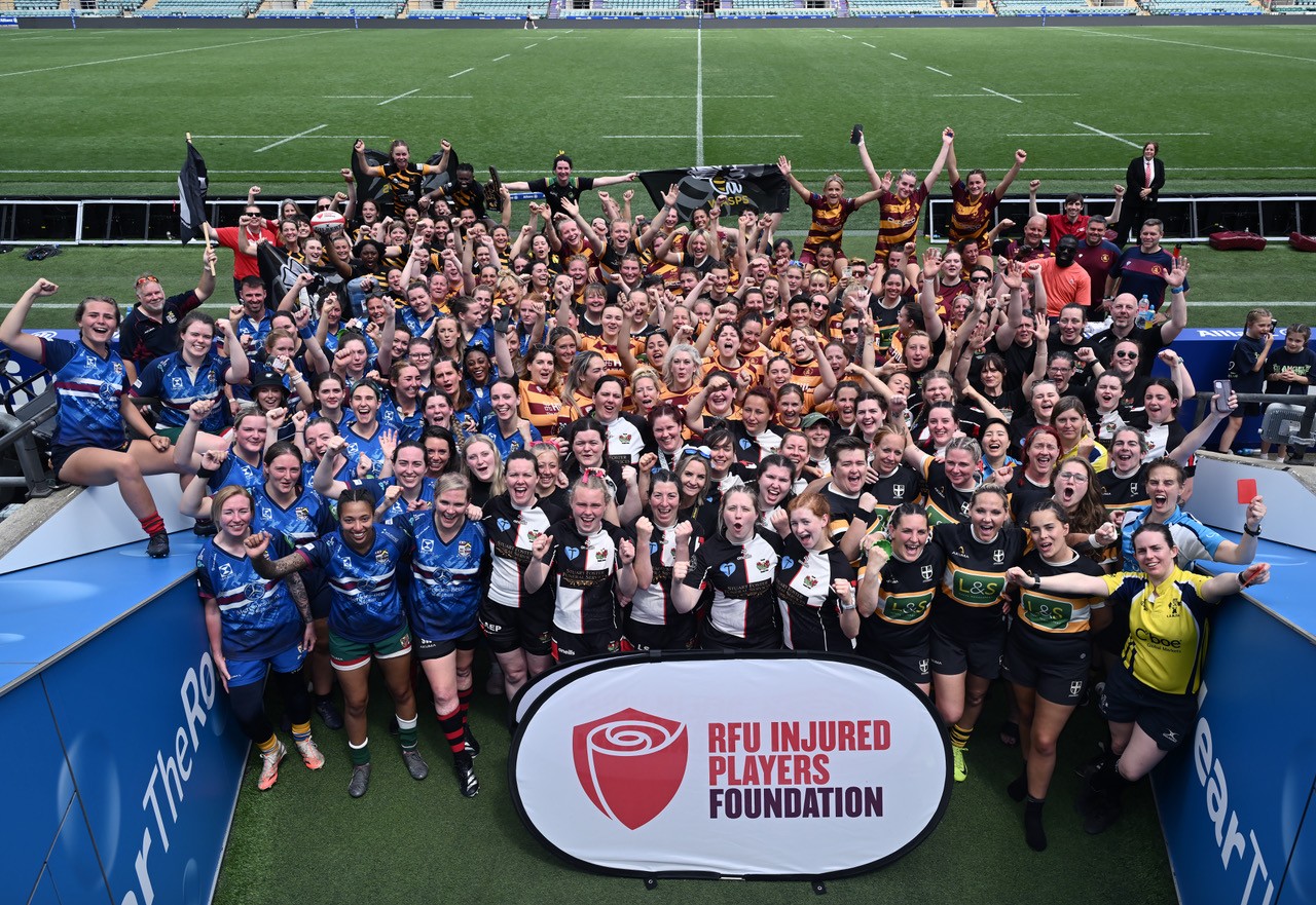 Image shows 100 female rugby players gathered together smiling with their hands up, whilst stood in the tunnel entrance to the Allianz Stadium, Twickenham pitch.